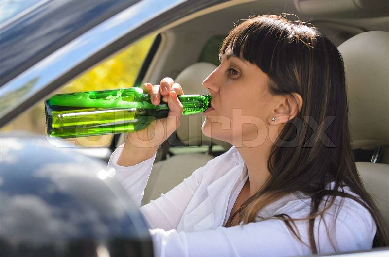 Alcoholic woman drinking and driving raising the bottle to her lips to take a swig as she steers the car, view through the side window, stock photo
