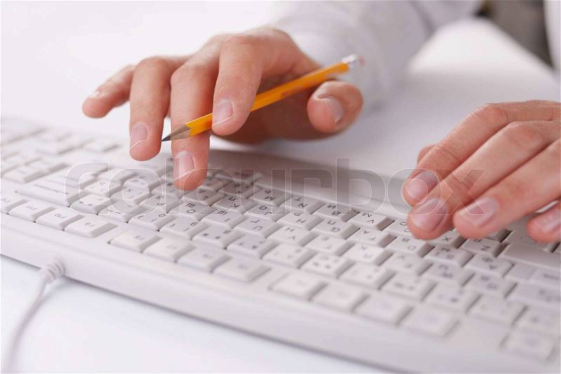 Man typing on a computer keyboard at work holding a pencil in the fingers of one hand as he enters data, close up view, stock photo