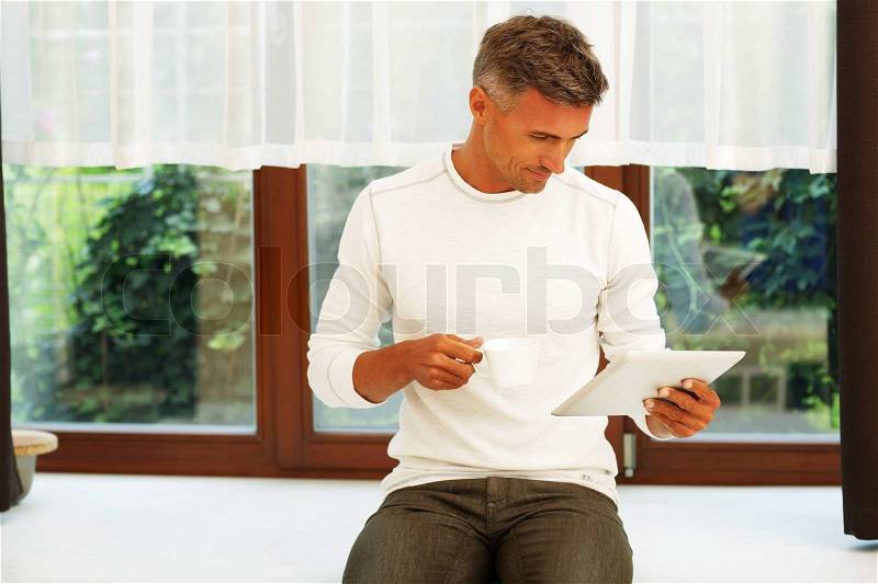 Happy young man at home drinking coffee and reading news on his electronic tablet, stock photo