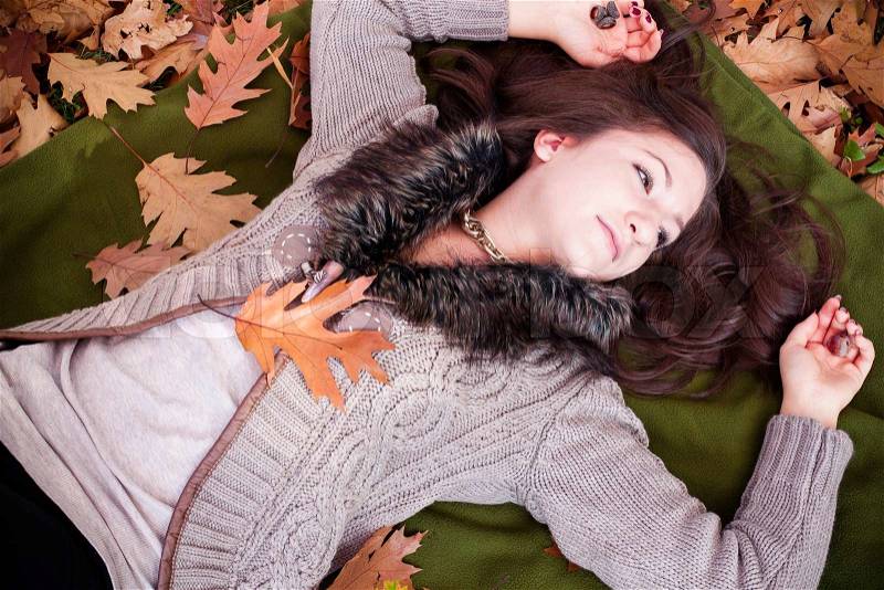 Attractive female laying down on the ground covered dry autumnal foliage in beautiful park and looking up in sky, stock photo