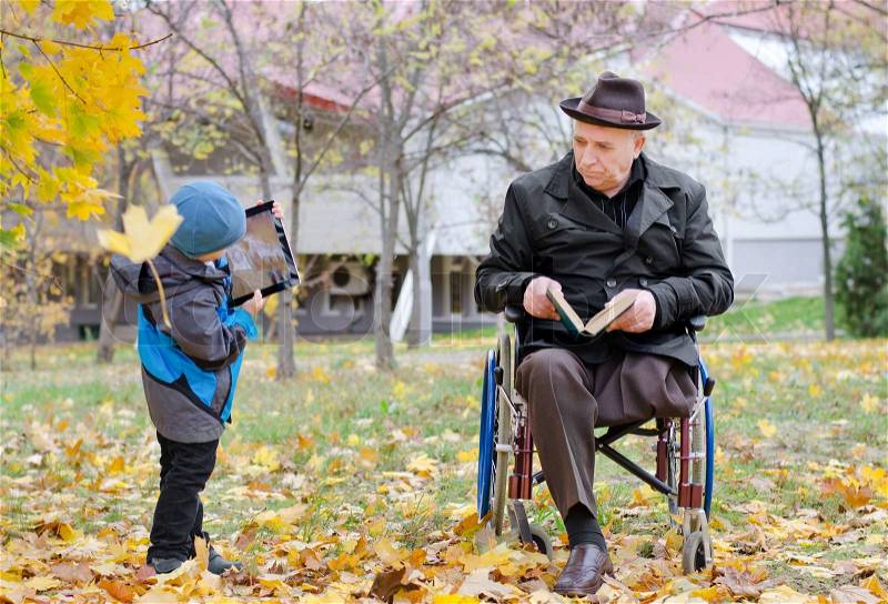 Disabled grandfather sitting in a wheelchair watching his young grandson playing with a tablet computer as the two enjoy a relaxing day in an autumn park together, stock photo