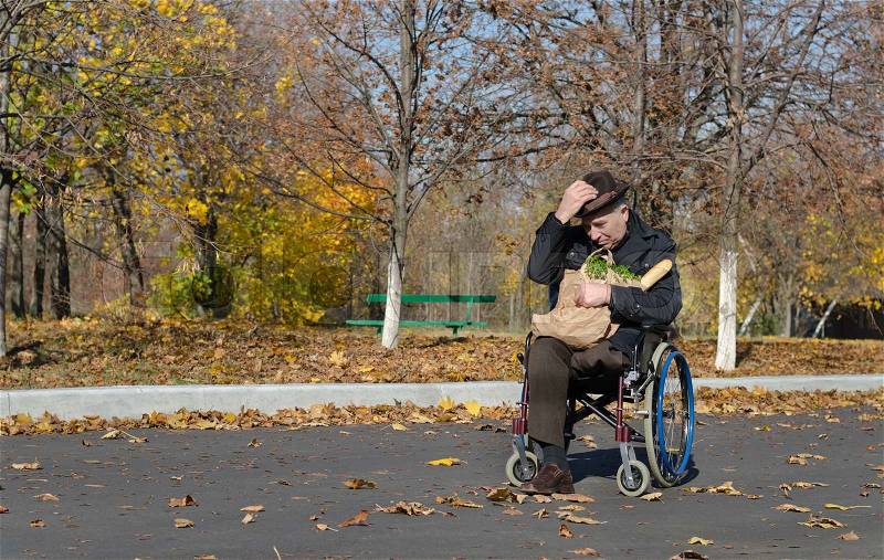Lonely handicapped man in a wheelchair parked in the street with a bag of groceries on his lap waiting for someone to come and assist him, stock photo