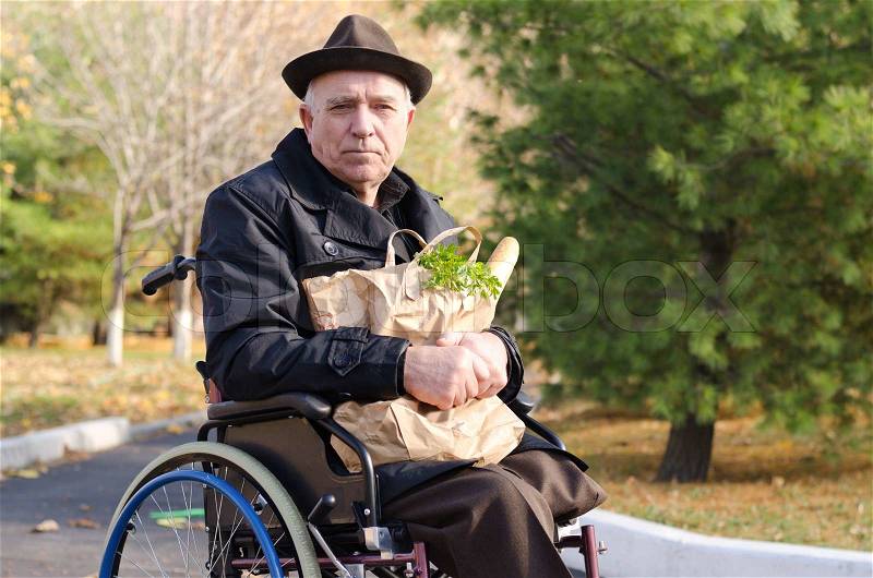 Senior handicapped man doing his grocery shopping sitting in the road in his wheelchair clutching a large bag of food and looking at the camera, stock photo