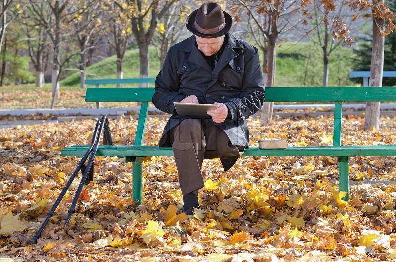 Elderly handicapped man on crutches sitting on a wooden bench dressed in a warm overcoat against the chilly autumn weather using a tablet computer in the park, stock photo