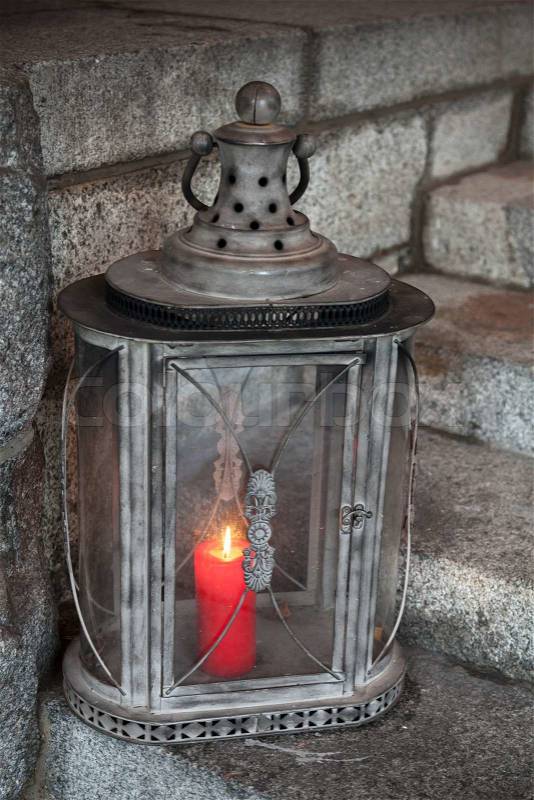 Old metal outdoor lamp with red burning candle stands on stone stairs, stock photo