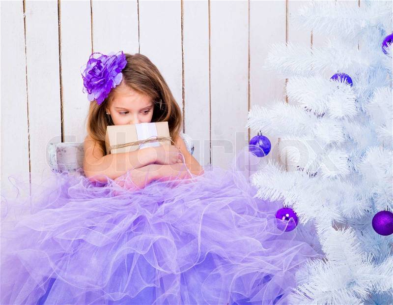 Sad little girl with a gift in hands near white Christmas tree, stock photo