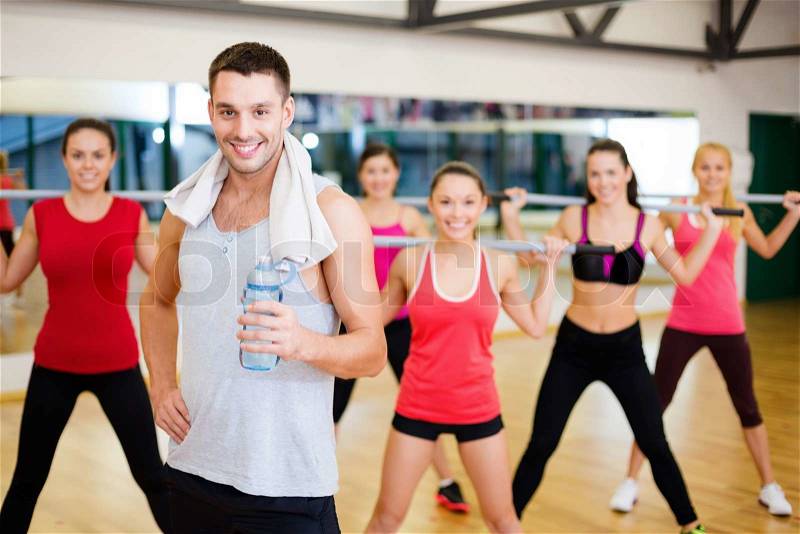 Fitness, sport, training, gym and lifestyle concept - smiling trainer in front of group of people working out with barbells in the gym, stock photo