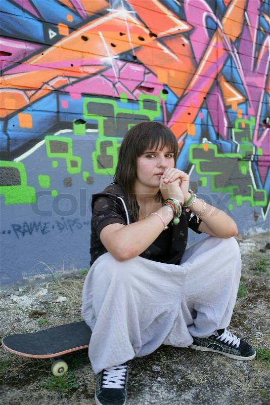Portrait of a girl sitting on a skateboard in front of a wall of graffiti, stock photo