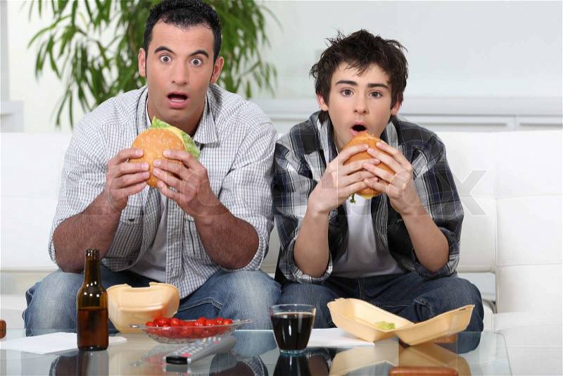 Father and son sat on the sofa eating burgers, stock photo