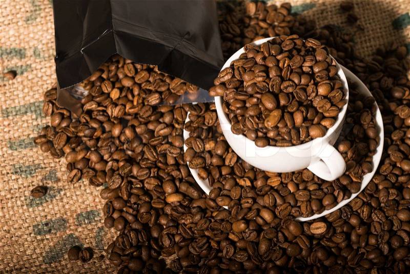 White coffee cup full of beans, surround by more coffee beans and a black bag, stock photo