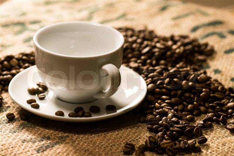 White coffee cup on a coffee sack with roasted beans around, stock photo