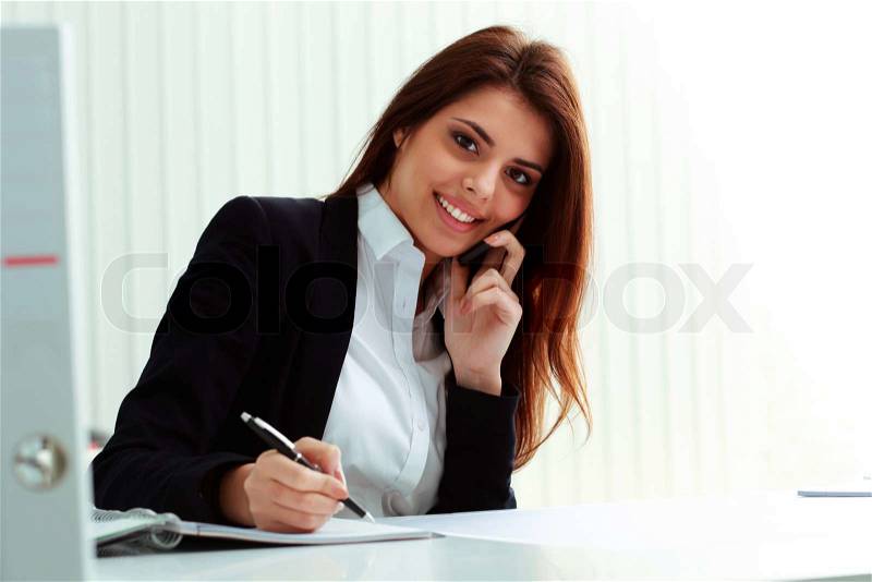 Young happy businesswoman talking on the phone and writing notes in office, stock photo
