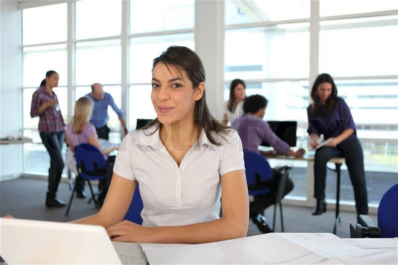 Smiling woman using a laptop in a busy office, stock photo