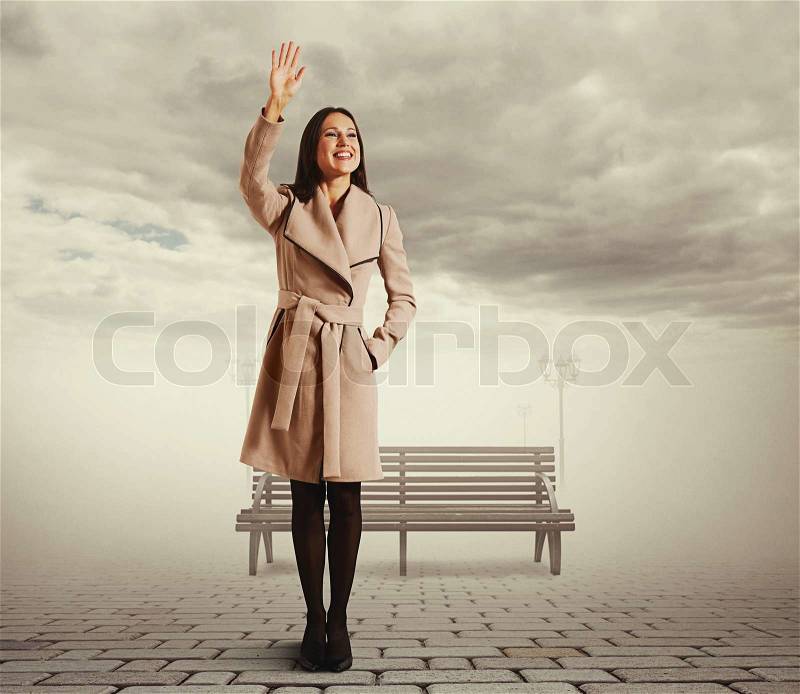 Happy young woman standing against bench and waving hand, stock photo