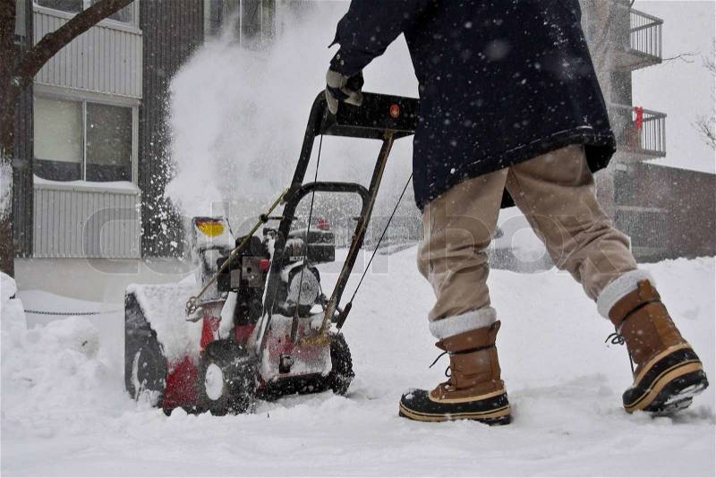 Man with a red snow blower trying to open the access to the garage door, near the building wall, stock photo