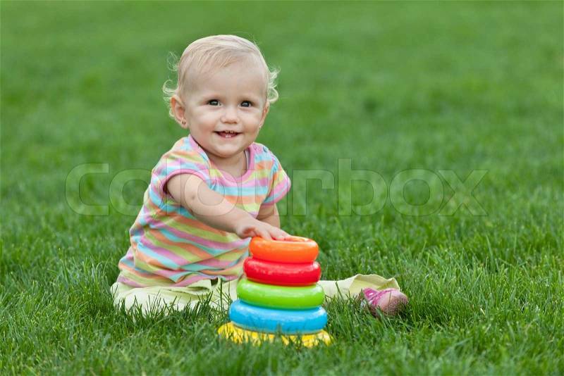 A pretty smiling little girl sitting on the green grass is playing with a toy pyramid , stock photo