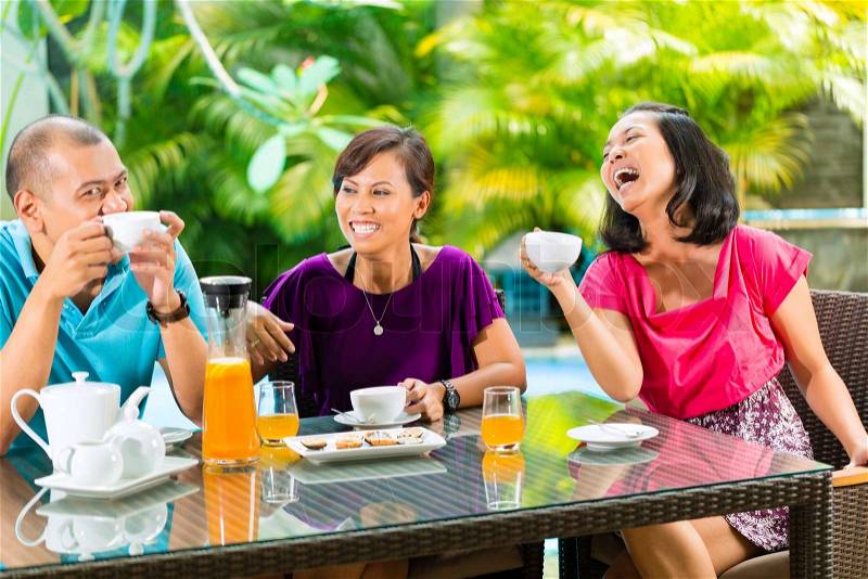Asian friends having coffee on the porch in front of a home, in the background a tropical garden, stock photo