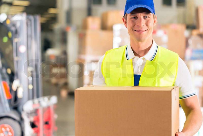 Warehouseman with protective vest holds package, he standing at warehouse of freight forwarding company, stock photo