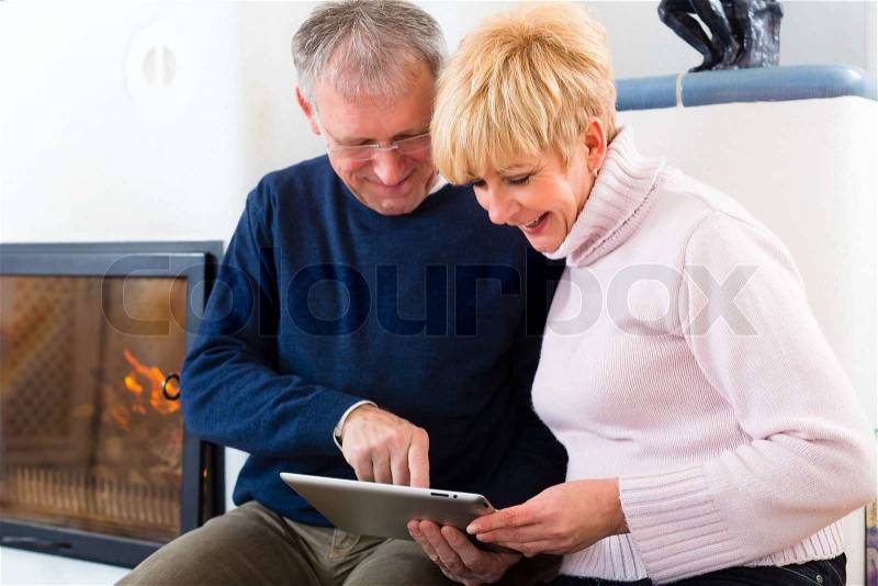 Quality of life - two elderly people sitting at home in front of the furnace, writing emails on the tablet computer, stock photo
