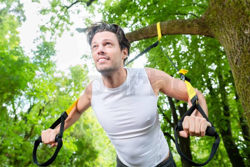 Man exercising with suspension trainer sling in City Park under summer trees for sport fitness, stock photo