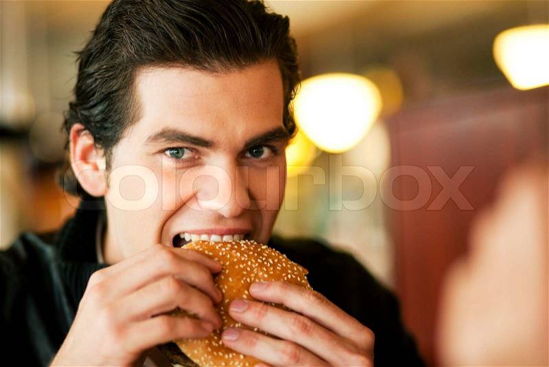 Man in a restaurant or diner eating a hamburger, he is hungry and having a good bite, shot with available light, very selective focus, stock photo