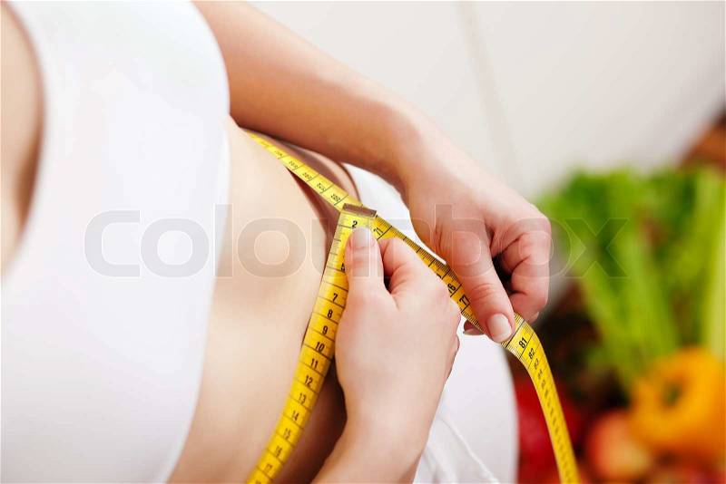 Thin and beautiful woman (only torso) measuring her waist with a tape measure, in the background fruit in a bowl, stock photo