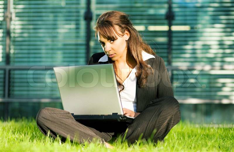 Female professional with a laptop on the lawn in front of a glass and steel facade, stock photo