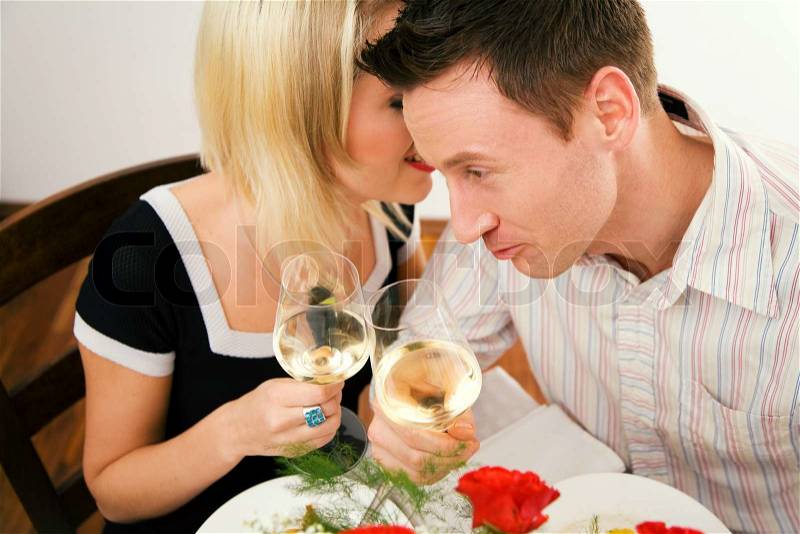 Young couple romantic dinner: both holding white whine glasses, she is whispering something; focus on his face, stock photo
