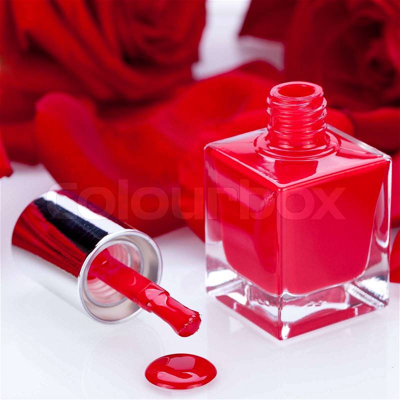Elegant red nail varnish in a stylish bottle surrounded by romantic red roses with the brush lying alongside the open bottle with a droplet on the white surface below, stock photo