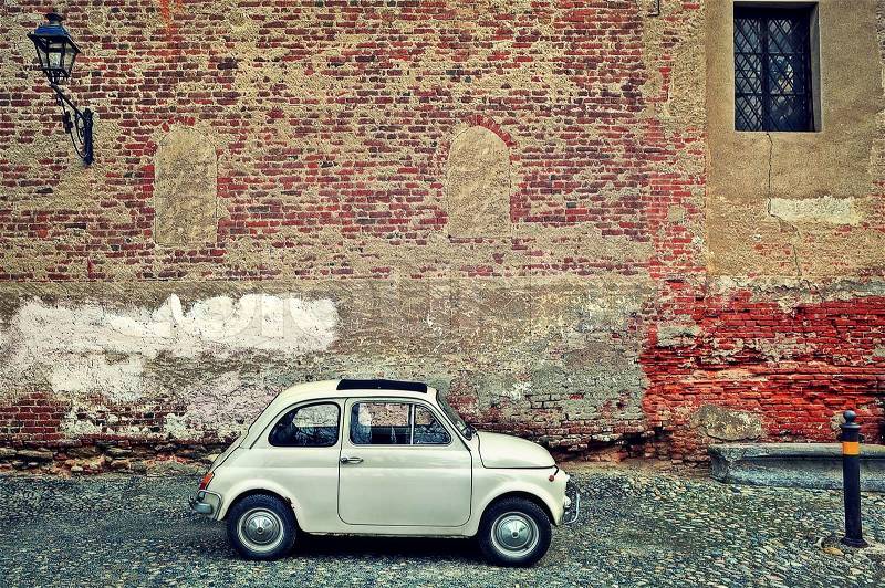 Small old car stand on cobbled street against red brick wall with lamppost in Saluzzo, Northern Italy, stock photo