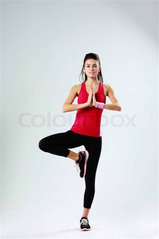 Young sport woman meditating while standing on one leg on gray background, stock photo
