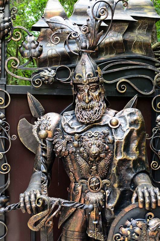 Ornate metal statue of a knight at arms wearing armour decorated with a lions head and dragon outdoors in a park or garden, stock photo