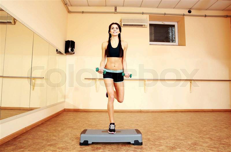Young cheerful fit woman doing exercises with dumbells on step board at gym, stock photo