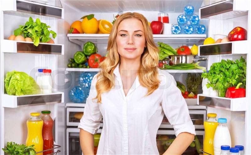 Portrait of cute serious female standing near open fridge full of healthy food, vegetables and fruits, healthy lifestyle concept, stock photo