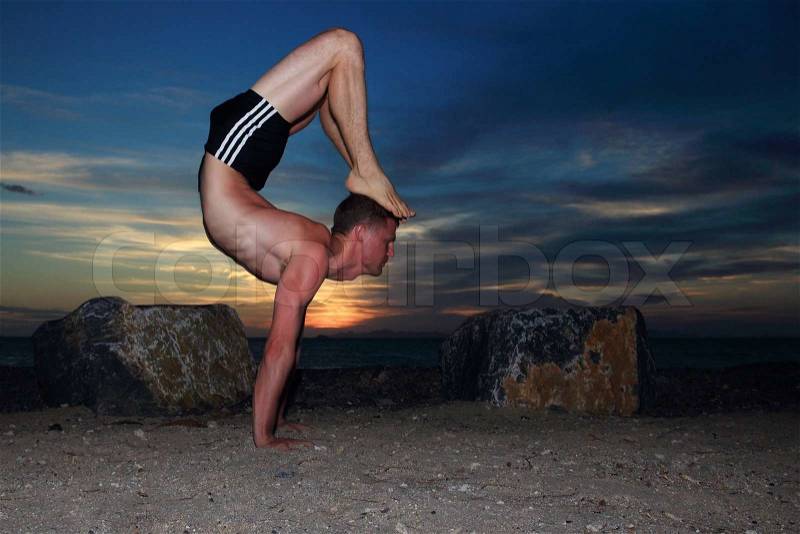 Man doing yoga Scorpion pose by the beach at dusk, stock photo