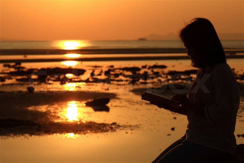 Silhouette woman reading book by beach at sunrise, stock photo