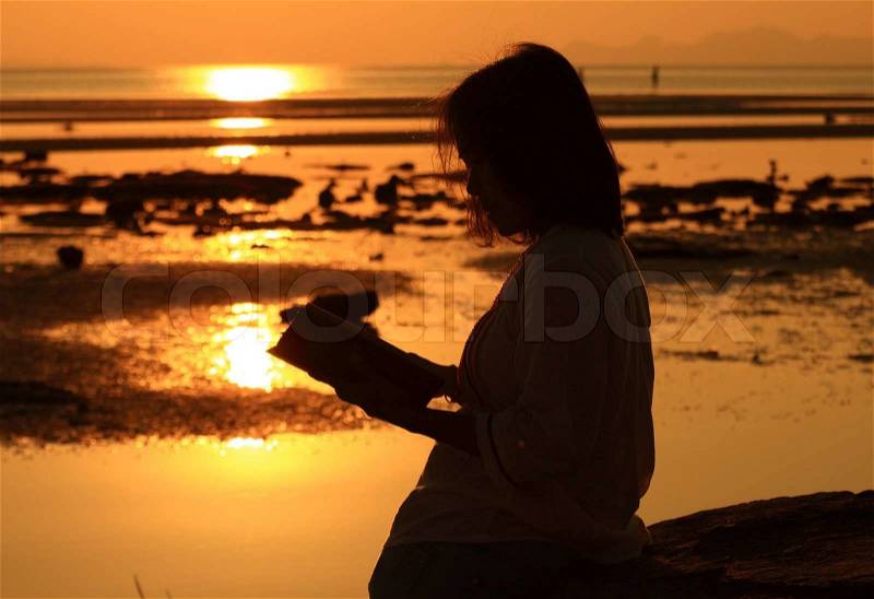 Silhouette woman reading book by beach at sunrise, stock photo
