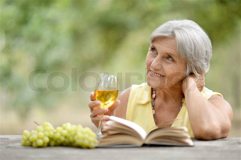 Senior woman reading book and drinking wine outdoors, stock photo