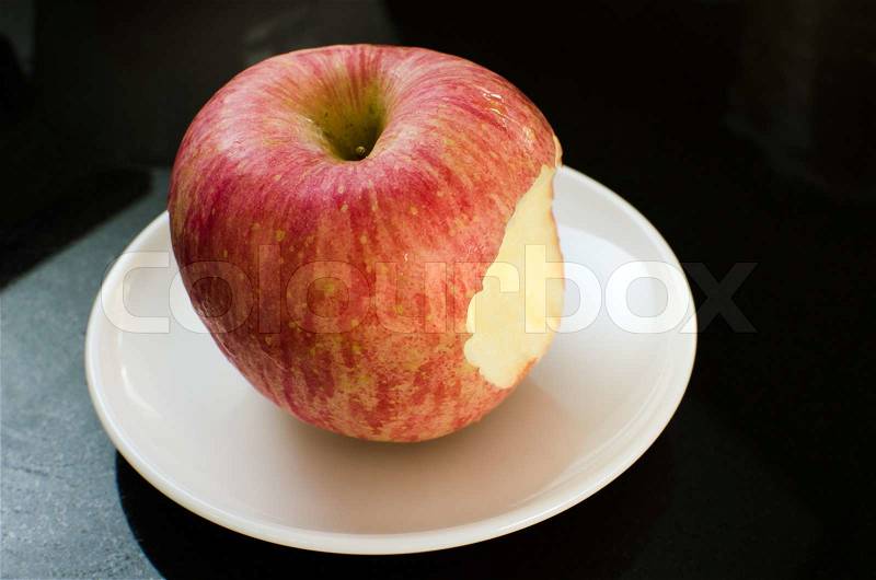 Big apple with bite taken in white ceramic dish on black granite table, stock photo
