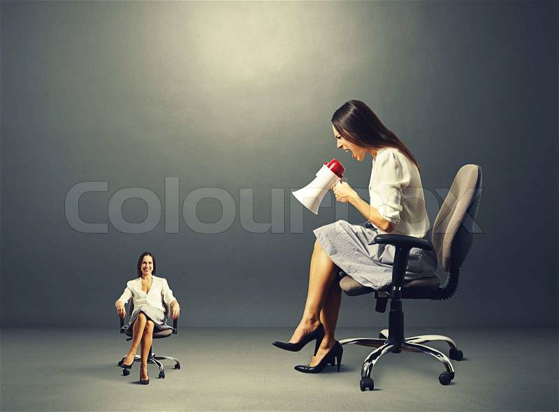 Angry young woman screaming at smiley calm woman on the chair over dark background, stock photo