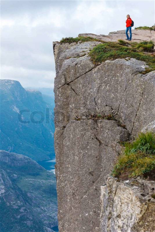 Woman hiker standing on cliff of Pulpit Rock / Preikestolen, Norway. Pulpit rock is a massive cliff 604 metres above Lysefjorden, almost flat, and is a famous tourist attraction in Norway, stock photo