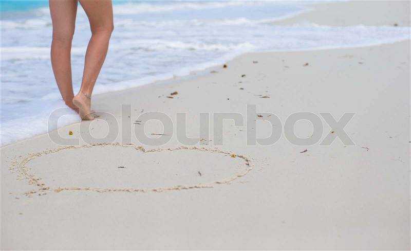 Women\'s beautiful smooth legs standing near big heart on white sand beach, stock photo
