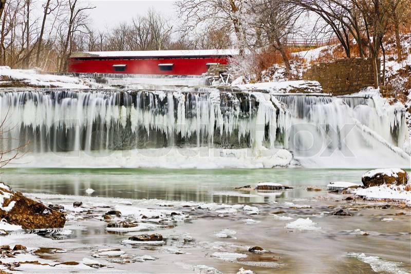 Whitewater flows over icicles at a frozen Upper Cataract Falls with a red covered bridge upstream. Photographed on Mill Creek near Cloverdale, Indiana, stock photo