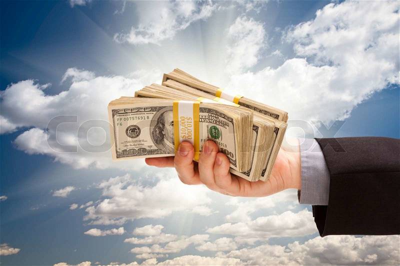 Male Hand Holding Stack of Cash Over Dramatic Clouds and Sky with Sun Rays, stock photo