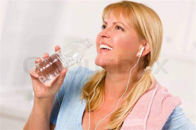 Pretty Blonde Woman with Towel and Ear Phones Drinking From Water Bottle in Her Kitchen, stock photo