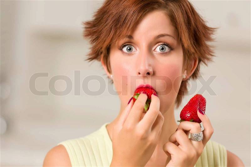 Pretty Wide-eyed Red Haired Woman Biting Strawberry in Her Kitchen, stock photo