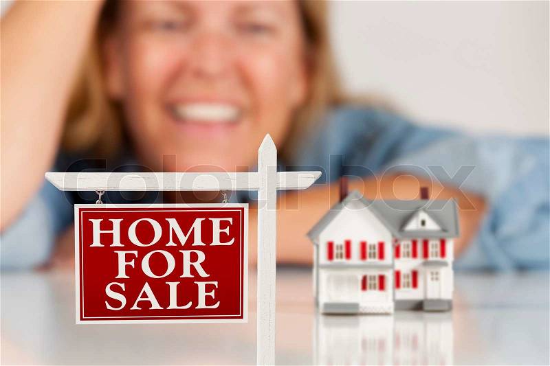 Smiling Woman Leaning on Hands Behind Home For Sale Real Estate Sign and Model House on a White Surface, stock photo