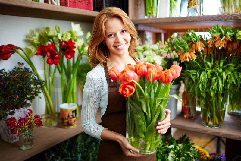 Portrait of young female florist with big vase of red tulips looking at camera, stock photo
